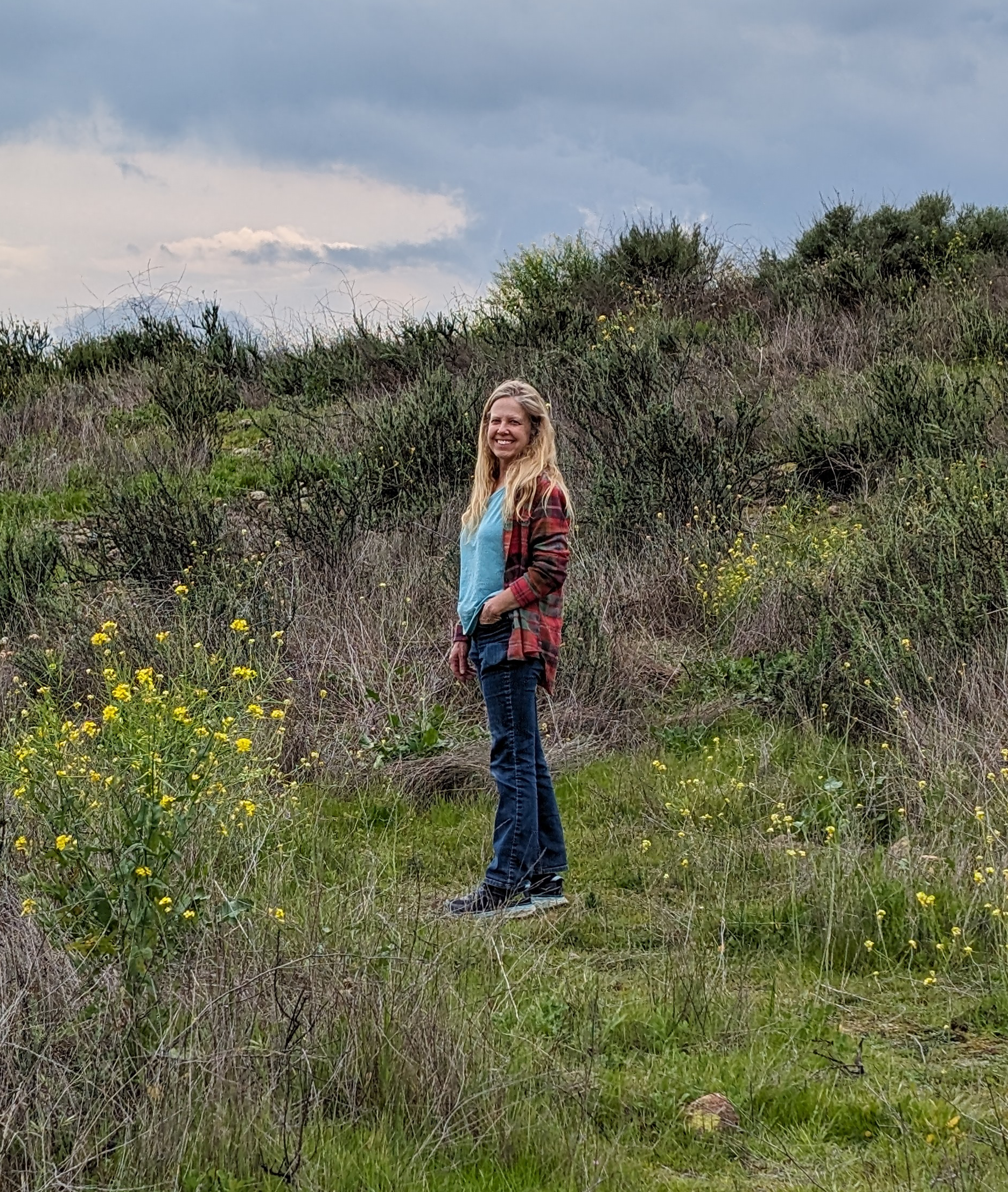 Shari Wasson stands on a green hillside, wearing a red flannel shirt with a blue top underneath. The sky above is overcast and lush grass extends across the hillside.